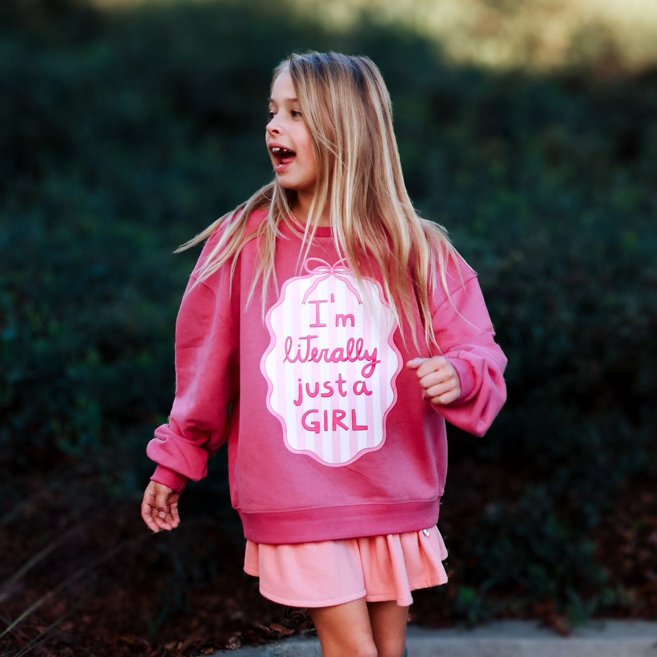 Young girl in a pink sweatshirt and skirt standing outdoors on a path with greenery.